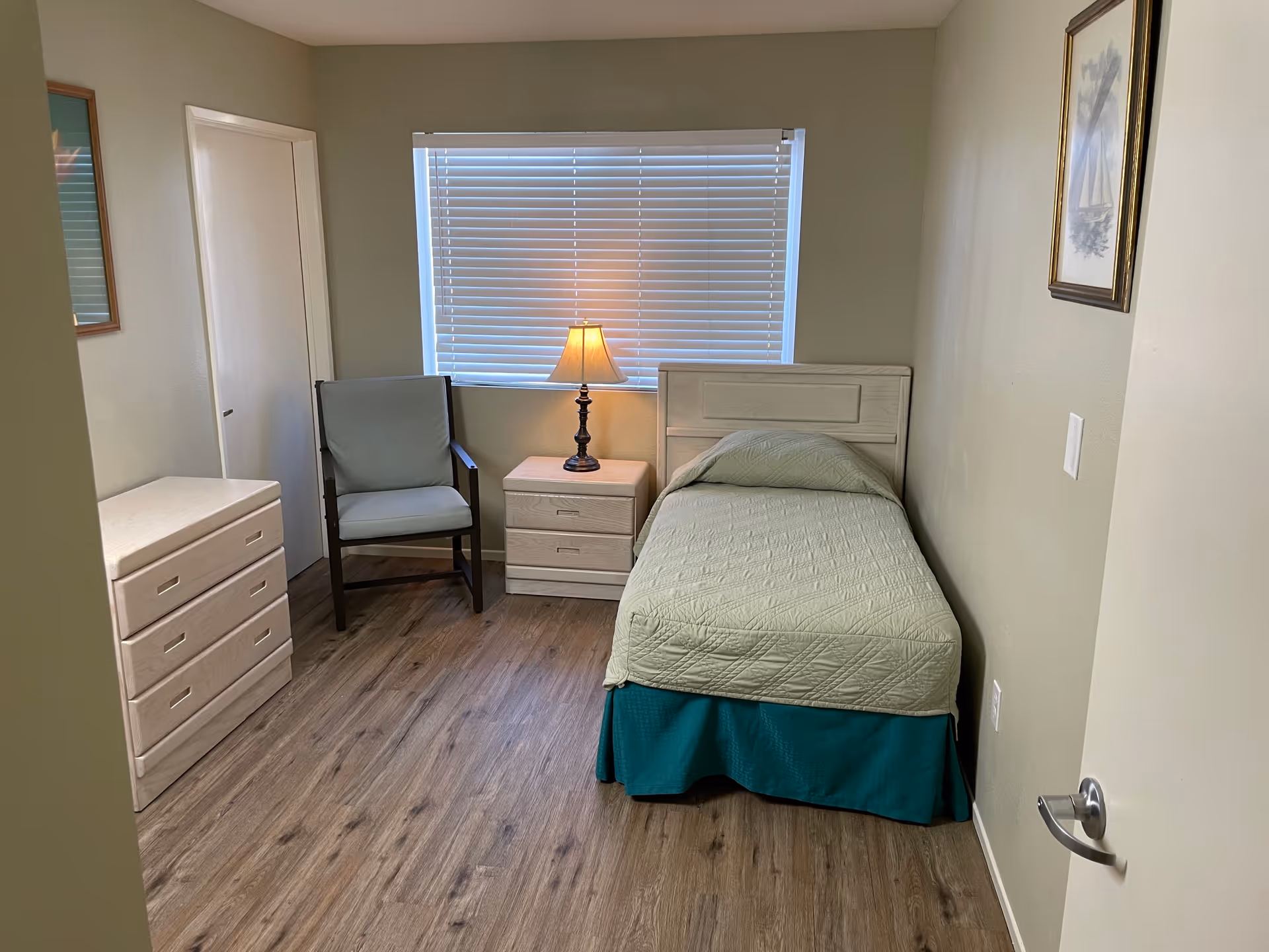 A small, simple bedroom with a single bed covered in a light green quilt and a teal bed skirt. Next to the bed is a wooden nightstand with two drawers and a lamp on top. There is a wooden armchair with a light blue cushion beside the nightstand. Across from the bed is a wooden dresser with three drawers. The room has light-colored walls, a window with closed blinds, and wood-look flooring. A framed picture hangs on the wall above the bed.