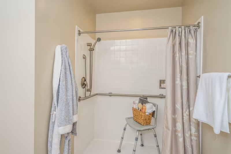 A bathroom shower area with white tiled walls and beige walls outside the shower. There is a metal grab bar along the back and side walls, a handheld showerhead, and a shower curtain with a floral pattern. A gray shower chair is inside the shower holding a basket with towels and toiletries. A gray bathrobe hangs on a hook outside the shower, and a white towel is draped over a towel bar on the right wall.