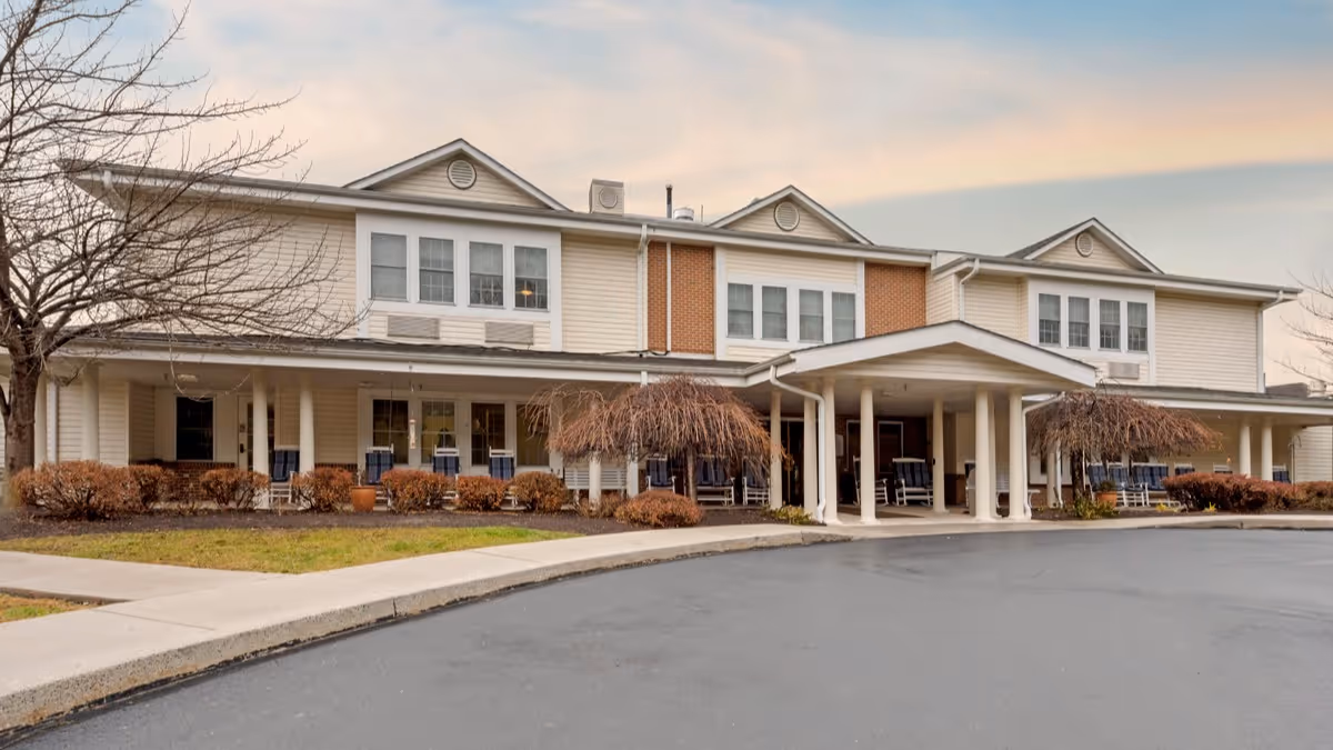 Front exterior of a two-story senior living facility with a covered porte-cochère, porch rocking chairs, and landscaped shrubs.