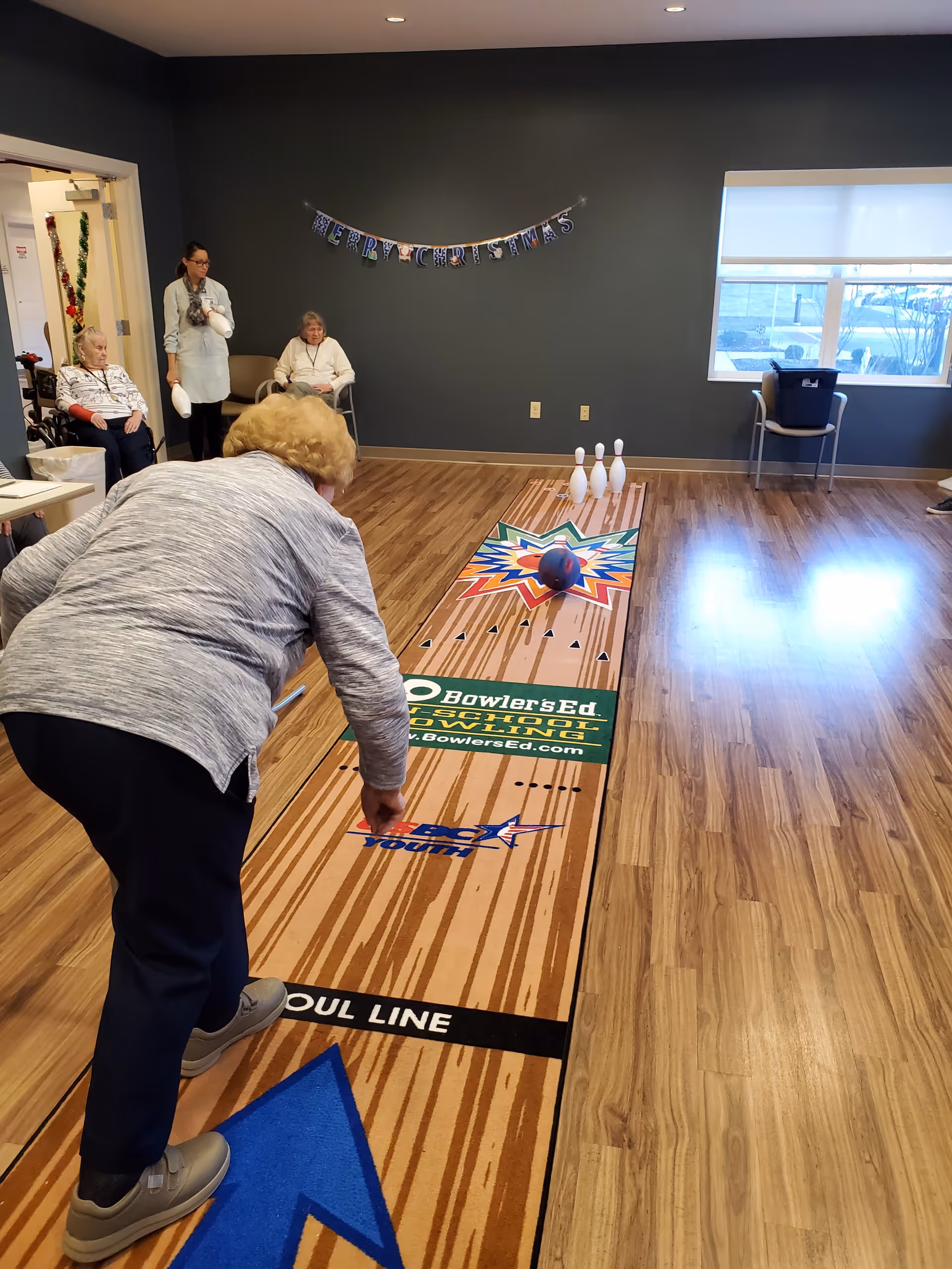 An elderly woman is playing indoor bowling on a wooden floor lane mat in a common room. Three bowling pins are set up at the end of the lane, and the woman is rolling a blue bowling ball towards them. In the background, three other elderly women are seated and watching. A 'Merry Christmas' banner is hanging on the dark gray wall, and a large window lets in natural light.