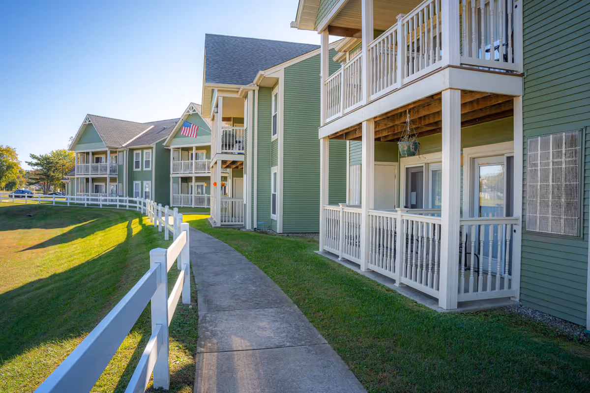 Exterior view of a senior living facility with green siding and white balconies. A concrete walkway runs alongside the building, bordered by a white fence and green grass. An American flag is visible hanging from one of the balconies.