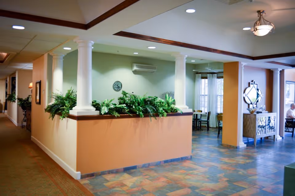 Interior view of a senior living facility showing a spacious common area with a tiled floor, beige and green walls, white columns, and a planter with green plants. There are tables and chairs in the background near windows with curtains, a decorative mirror on the wall, and a person seated at a table on the right side.