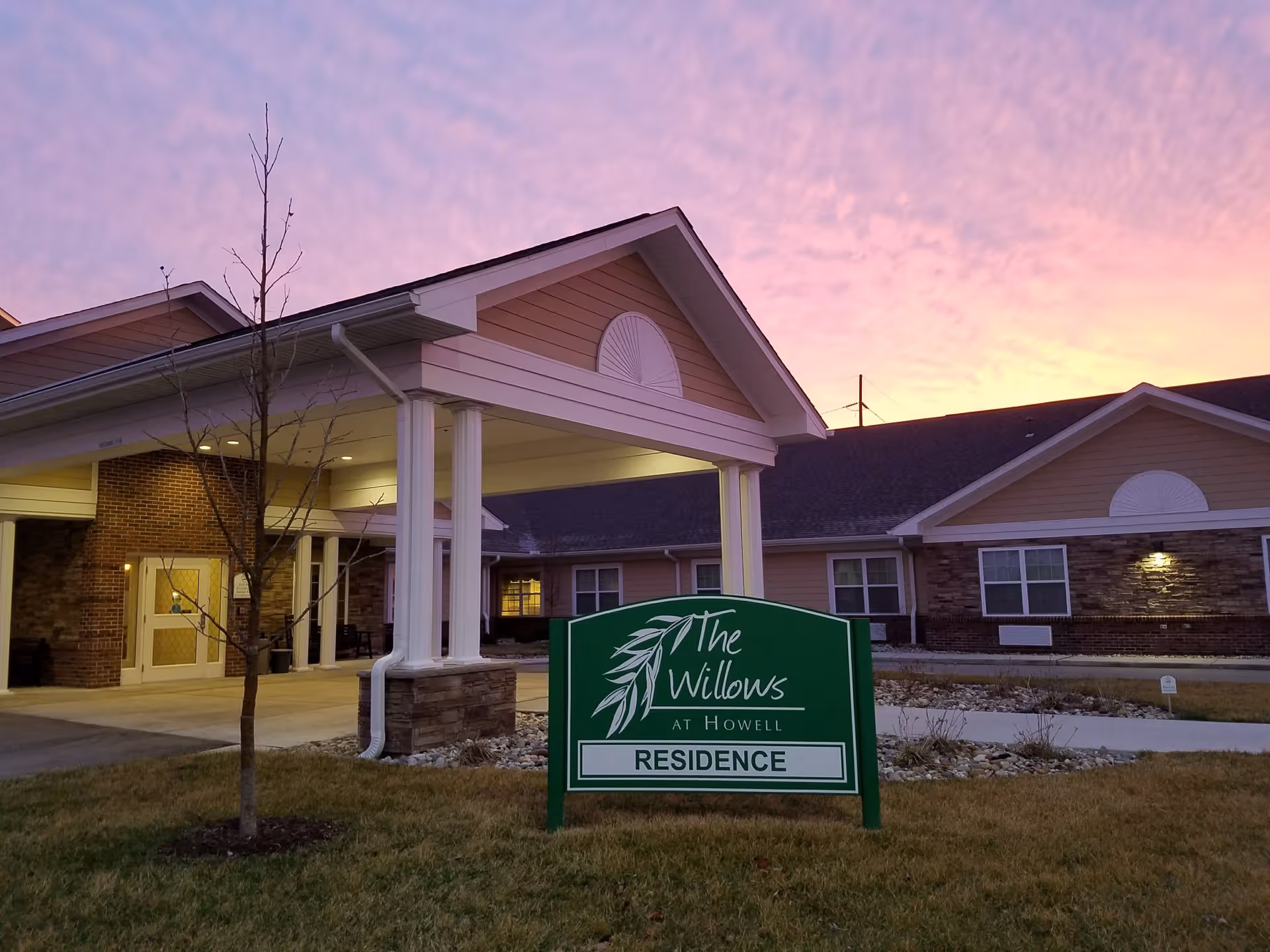 Exterior view of The Willows At Howell residence building at sunset, showing the entrance with white columns, brick and siding walls, a small tree in the foreground, and a green sign with the facility name.