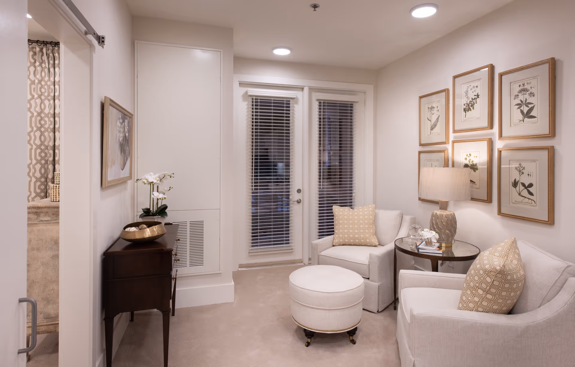 A cozy living room area with two beige armchairs, a round ottoman, and a small round glass-top side table with a lamp and decorative items. The walls are adorned with six framed botanical prints. There is a dark wooden console table with a decorative bowl and a white orchid plant. French doors with blinds lead outside, and a glimpse of a bathroom with a patterned shower curtain is visible through an open sliding door.