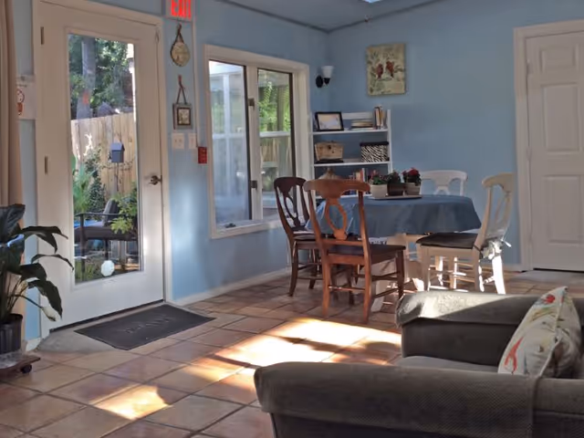 A cozy interior room with light blue walls and tiled floor, featuring a dining table covered with a blue tablecloth surrounded by mismatched wooden chairs. There is a gray upholstered armchair with a decorative pillow in the foreground. A glass door and window let in natural light and show a glimpse of an outdoor garden area. A small bookshelf with books and decorative items is against the wall.