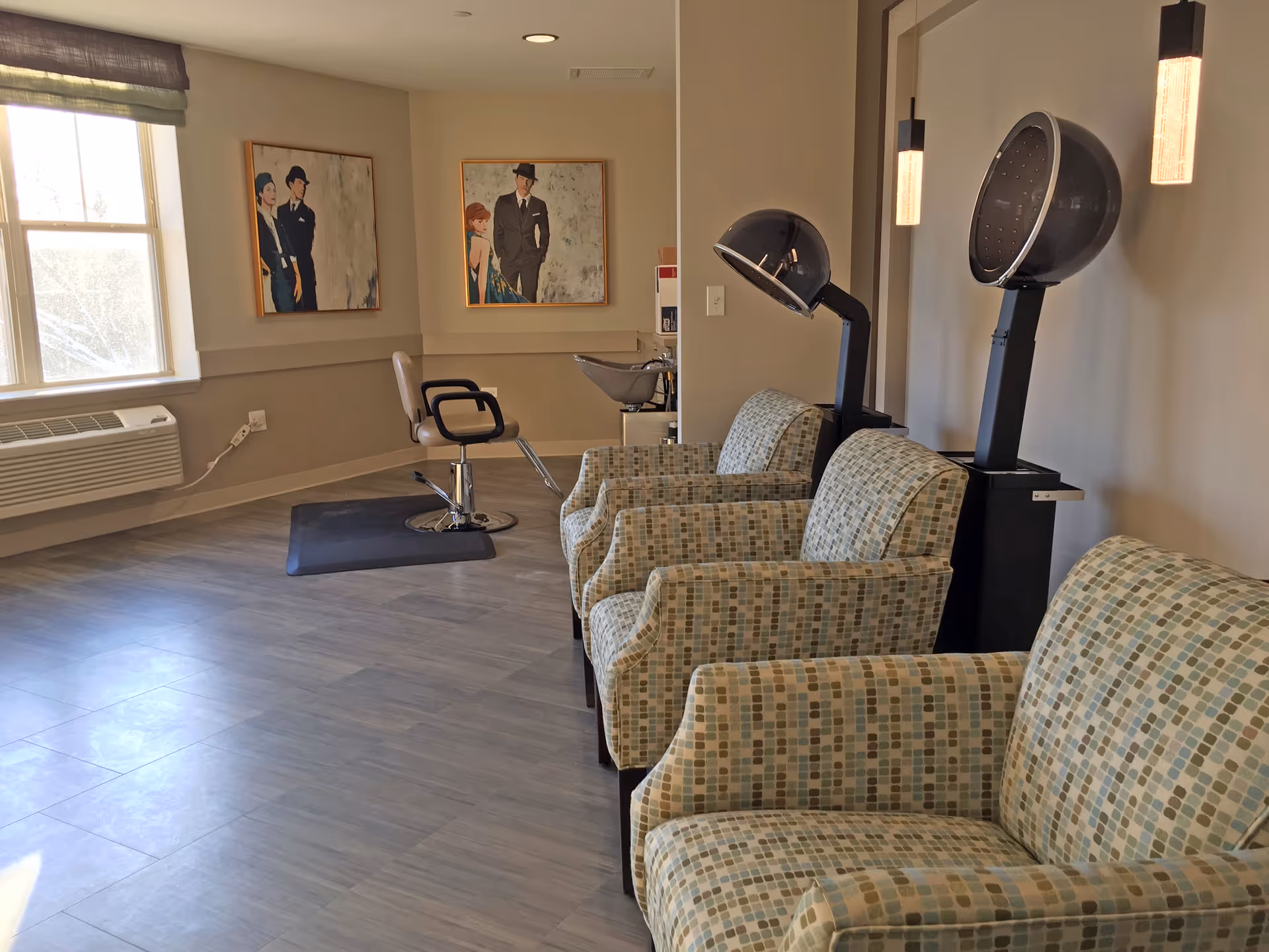 Interior of a salon area in a senior living facility with three patterned armchairs lined up next to two black hair drying stations. A single salon chair is positioned near a window with an air conditioning unit below it. Two framed paintings of people in vintage attire hang on the wall.