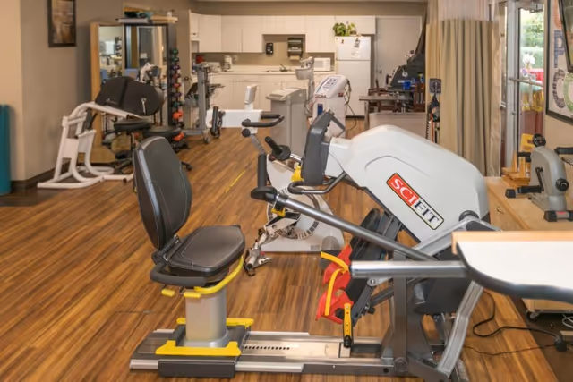 Interior view of a rehabilitation and healthcare facility gym area with exercise equipment including a SciFit machine, stationary bikes, and weight racks. The room has wood flooring, a kitchenette area in the background, and large windows with curtains allowing natural light.