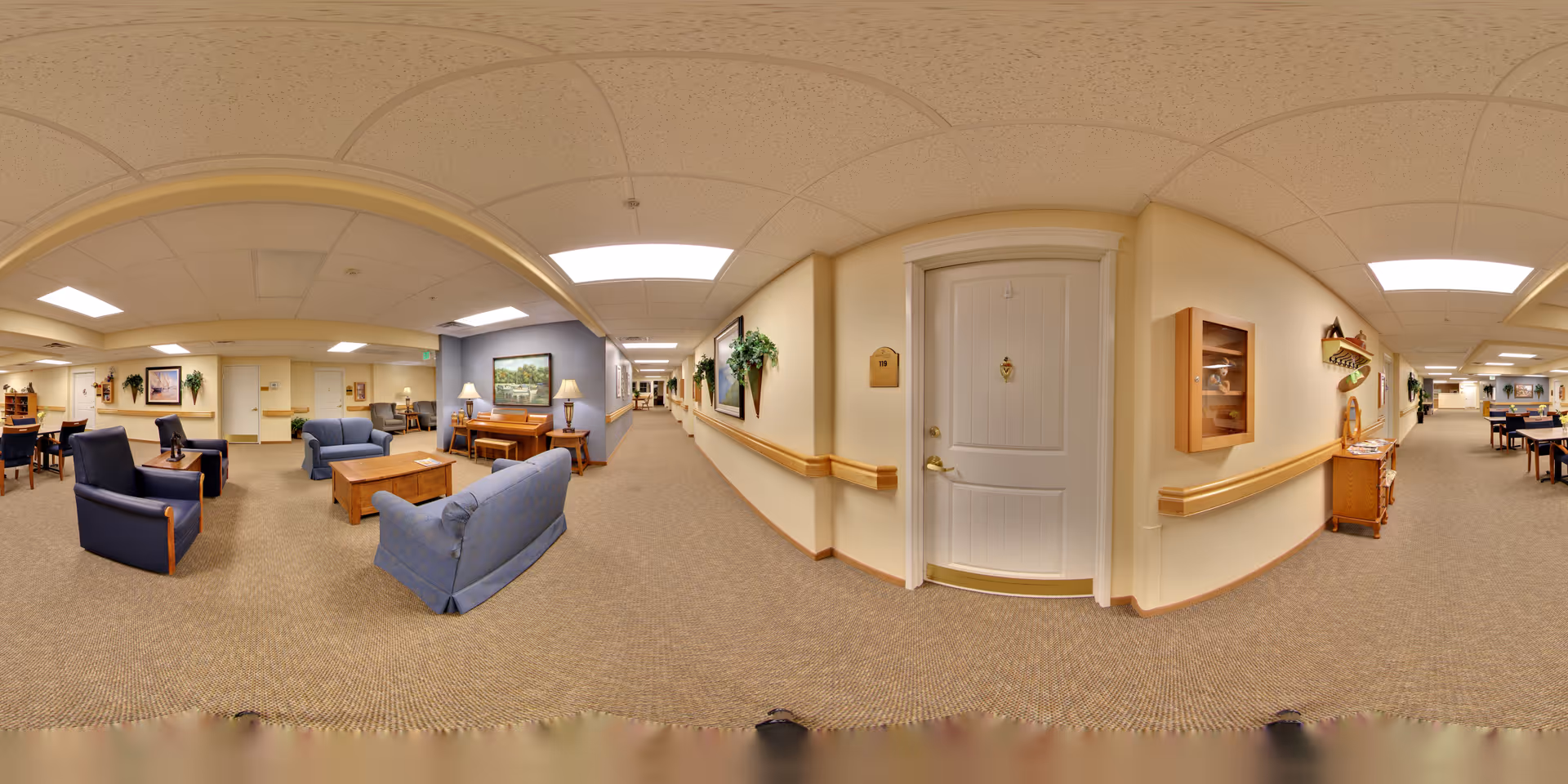 A wide-angle view of a senior living facility interior hallway with seating areas. The space features blue upholstered chairs and sofas arranged around wooden coffee tables. The walls are decorated with framed artwork and plants, and there is a white door with a brass handle and a plaque. The ceiling has recessed lighting and acoustic tiles, and the floor is carpeted in a neutral tone.