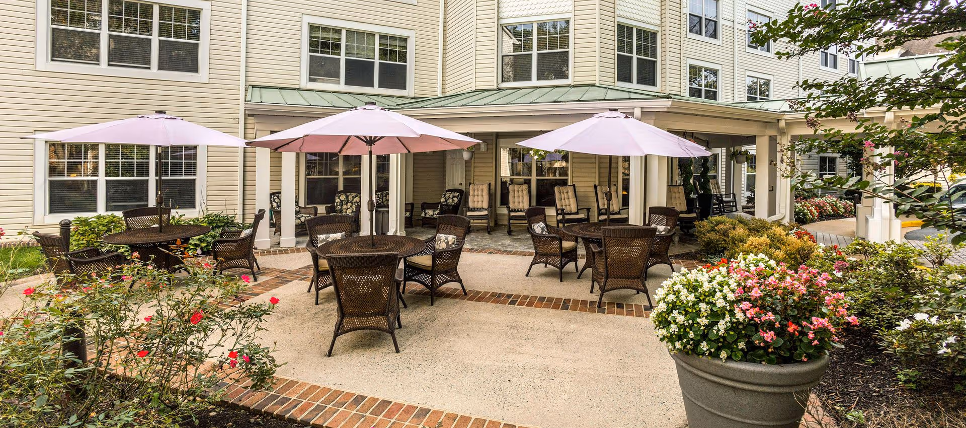 Outdoor patio area with round tables, umbrellas and wicker chairs in front of a multi-story building, surrounded by potted and planted flowers.