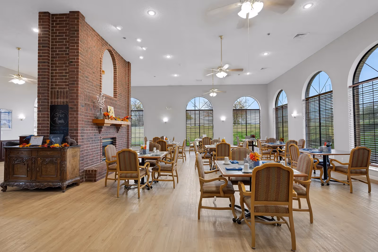 A spacious dining room with multiple tables and chairs arranged neatly. The room features large arched windows with blinds, allowing natural light to fill the space. There is a prominent brick fireplace in the center with a wooden mantel decorated with autumn-themed items. Ceiling fans and recessed lighting illuminate the room, and a wooden cabinet with a chalkboard sign reading 'Give Thanks' is visible on the left side.