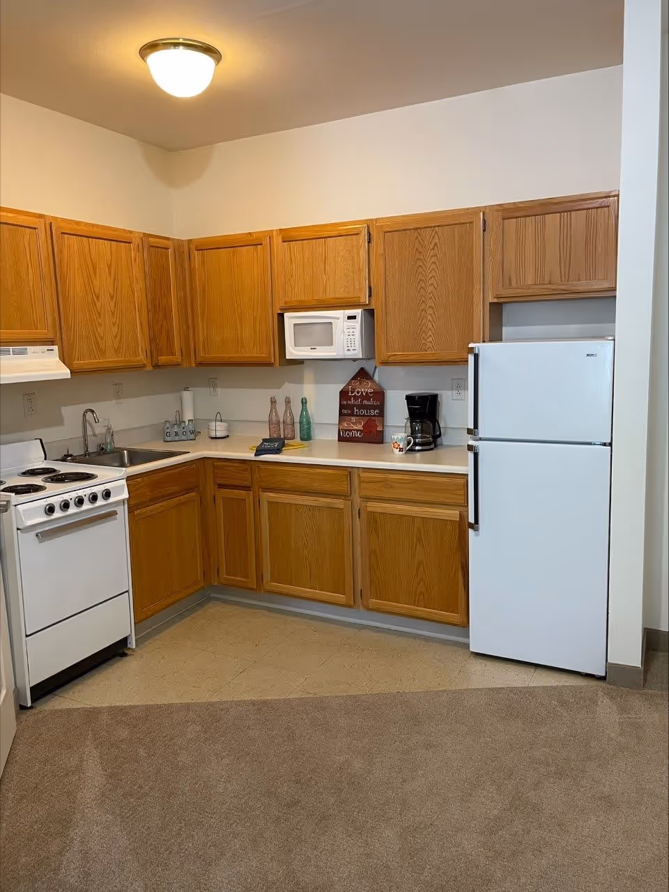 A small kitchen area with wooden cabinets, a white electric stove, a white microwave, a white refrigerator, a coffee maker, and various decorative items on the countertop including bottles and a sign that reads 'Love is what makes our house a home'. The floor transitions from carpet to linoleum near the kitchen.