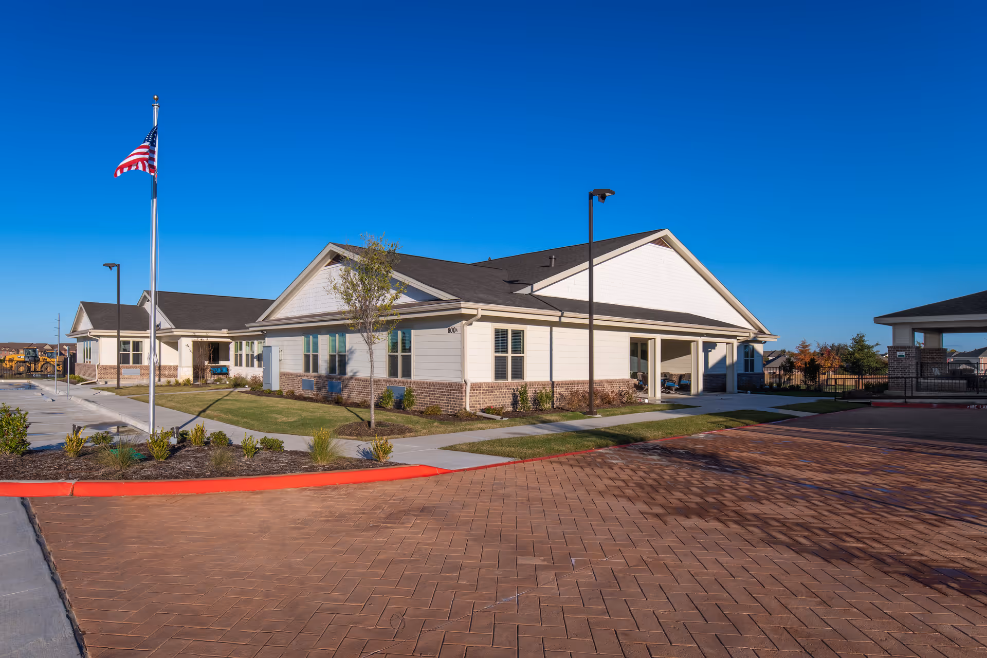 Exterior view of a single-story assisted living facility building with white siding and brick accents under a clear blue sky. There is a paved driveway and sidewalk leading to the entrance, a flagpole with an American flag, and some landscaping with small plants and trees.