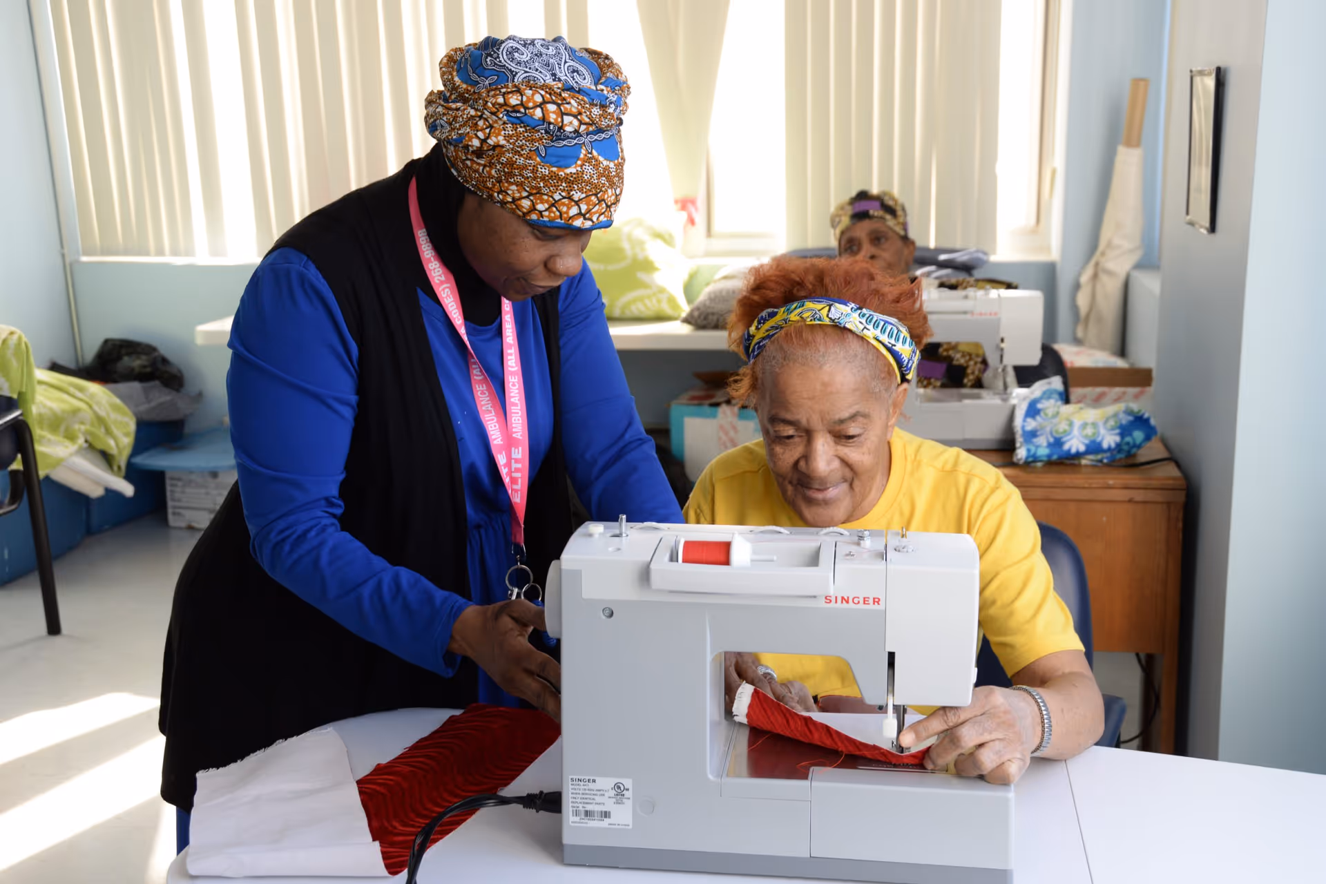 An elderly woman in a yellow shirt is using a sewing machine with the assistance of a caregiver wearing a blue outfit and a patterned headscarf. They are in a well-lit room with windows covered by vertical blinds. Another elderly woman is visible in the background, also working with a sewing machine.