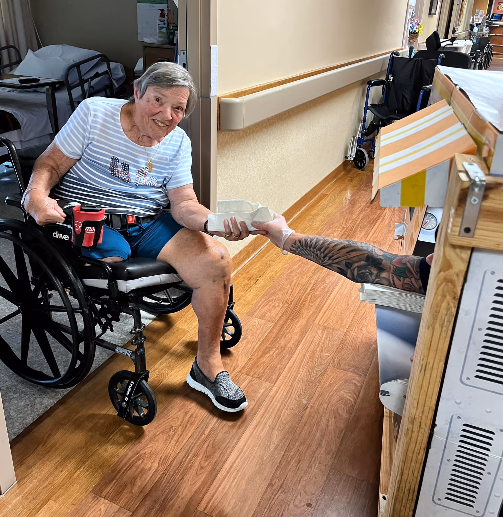 An elderly woman in a wheelchair is smiling and reaching out to receive a small white container from a person with a tattooed arm behind a wooden counter in a hallway with wood flooring and beige walls. There are other wheelchairs and beds visible in the background.