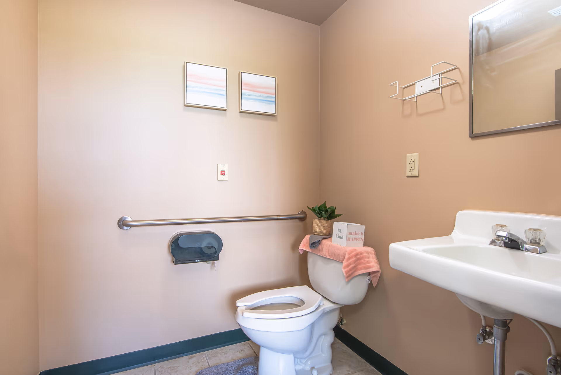 A small bathroom with beige walls featuring a white toilet with the lid open, a pink towel and a small potted plant on top of the toilet tank. There is a white sink with a mirror above it on the right side. Two framed abstract pictures hang on the wall above a metal grab bar and a toilet paper dispenser. The floor is tiled and there is a gray bath mat in front of the toilet.