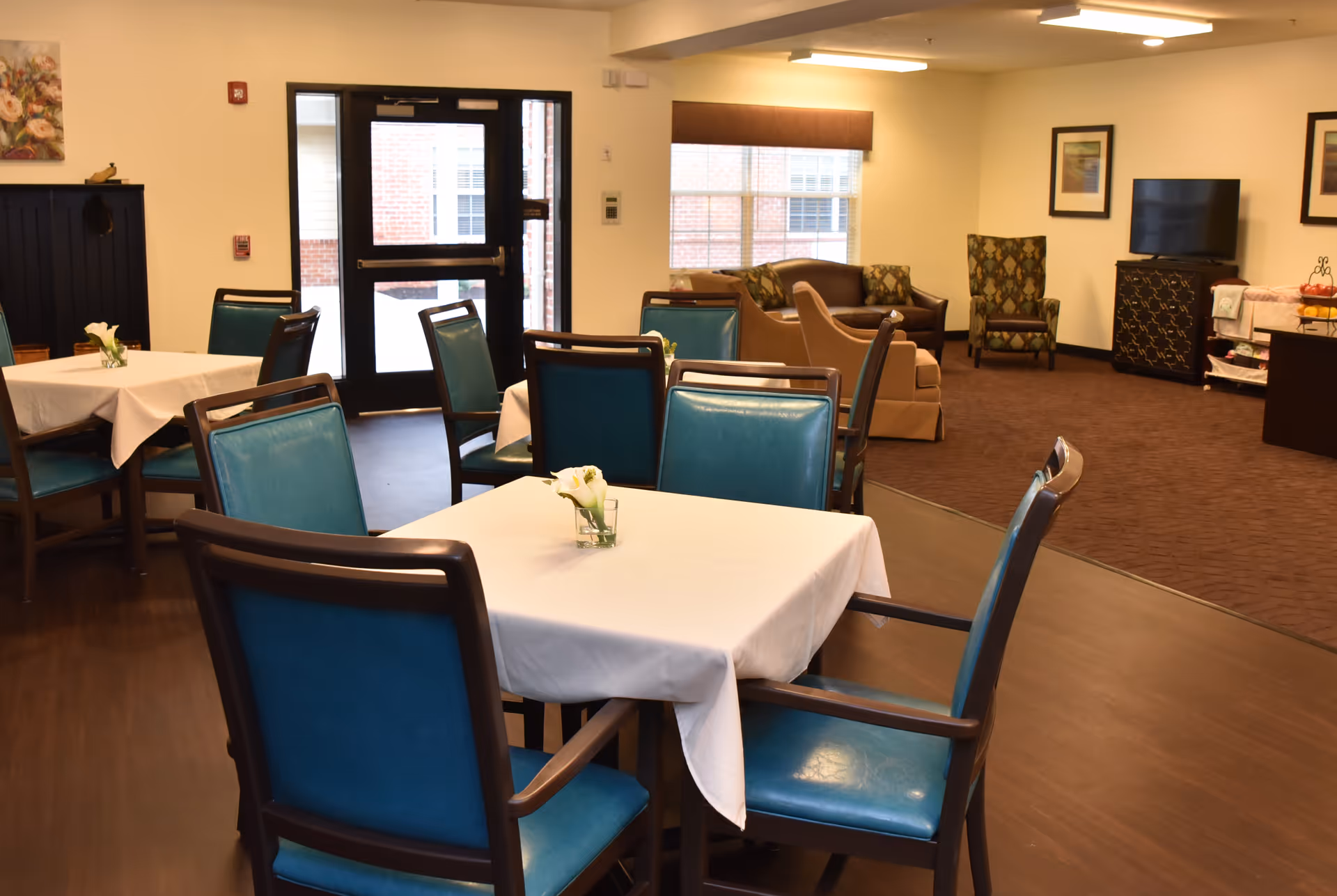 Dining room with square tables covered in white tablecloths, teal chairs, and a lounge area with couches and a TV.