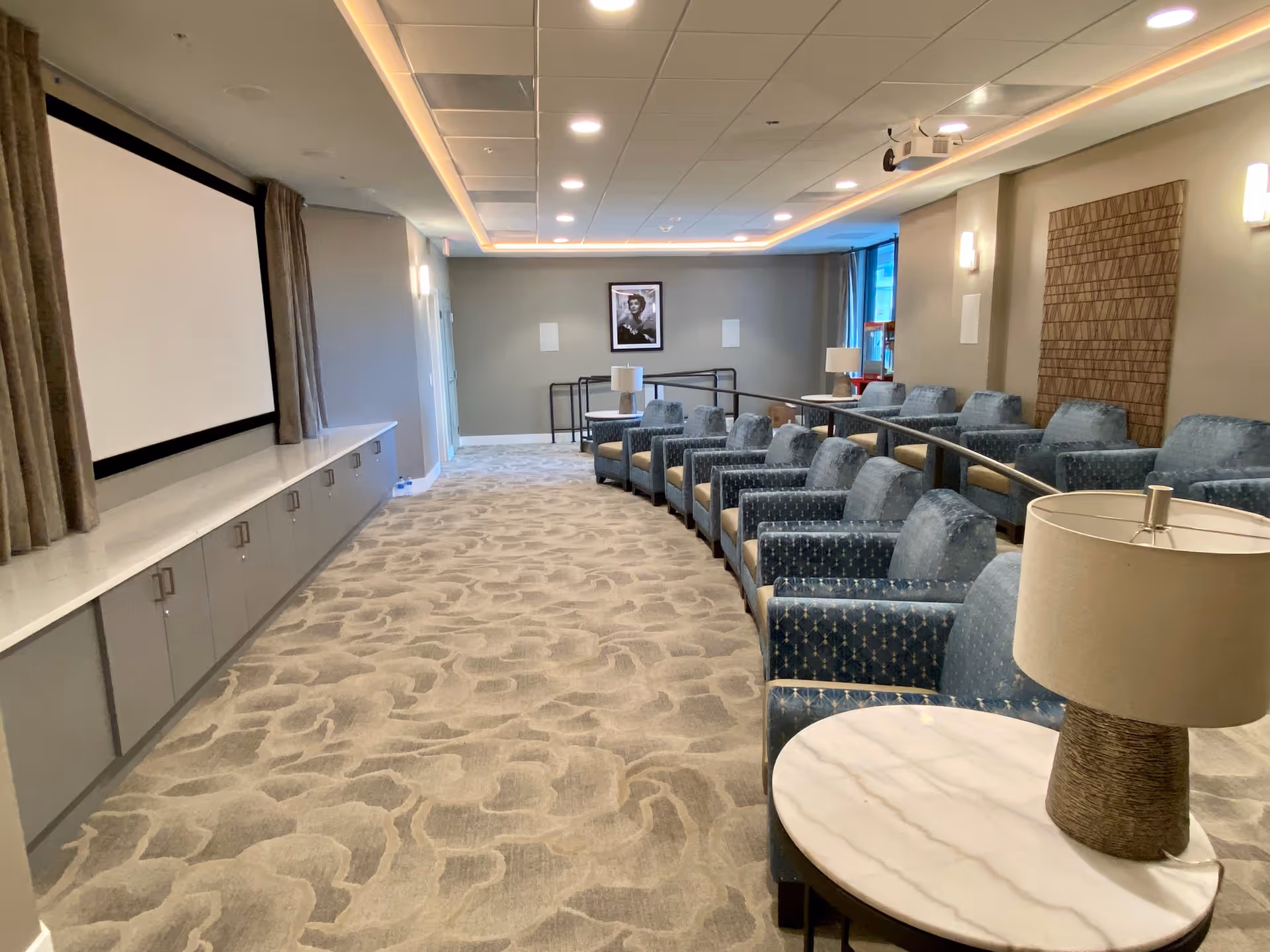 A modern theater room with rows of blue upholstered armchairs arranged in a tiered seating layout facing a large white projection screen. The room has beige walls, patterned carpet, ceiling lights, and a framed black and white photograph on the far wall. There are small tables with lamps beside some chairs.