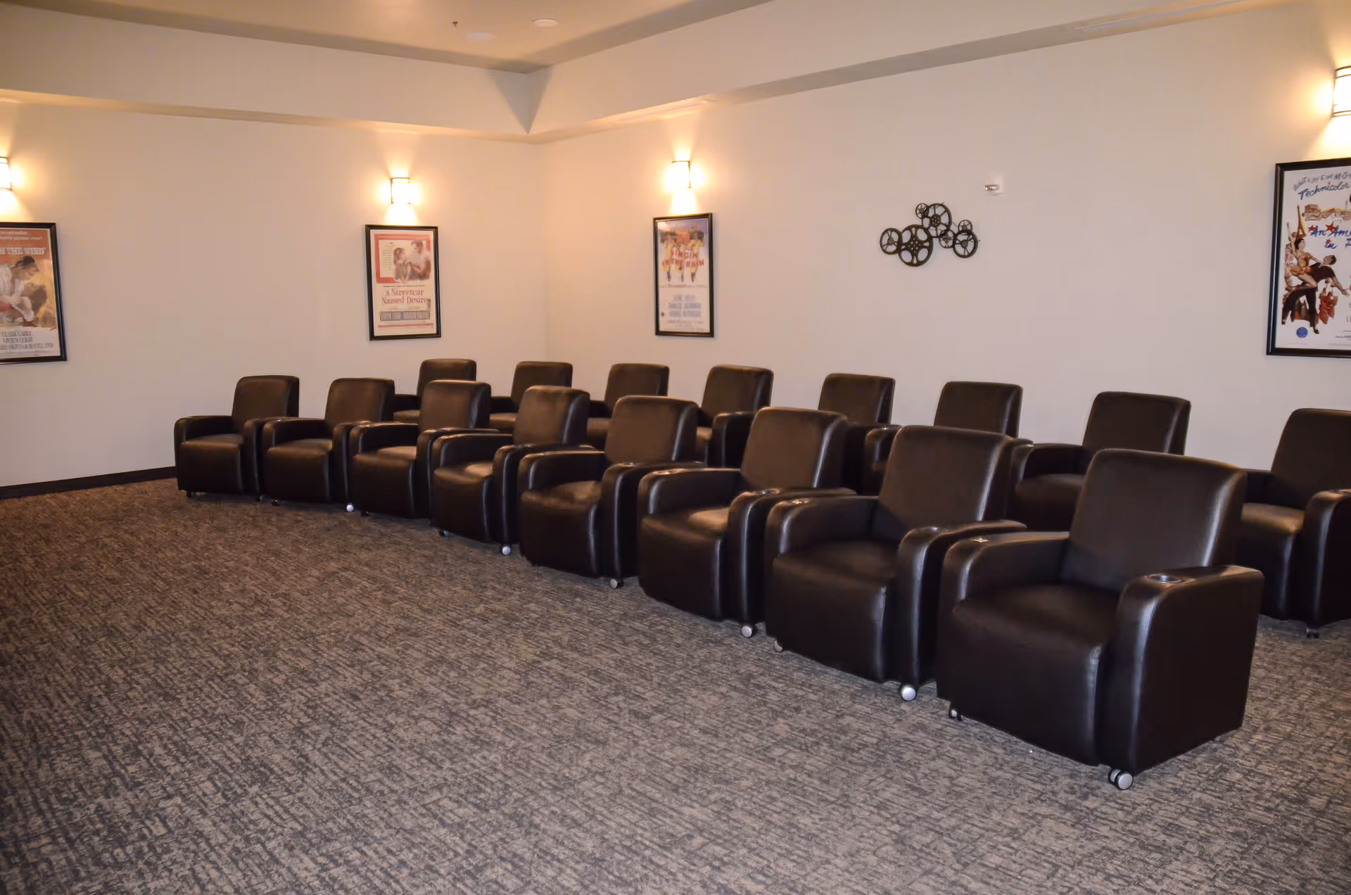 A room with two rows of black leather recliner chairs arranged facing forward, resembling a small theater or media room. The walls are light-colored with framed movie posters and decorative wall art. The floor is carpeted in a patterned design, and wall sconces provide soft lighting.