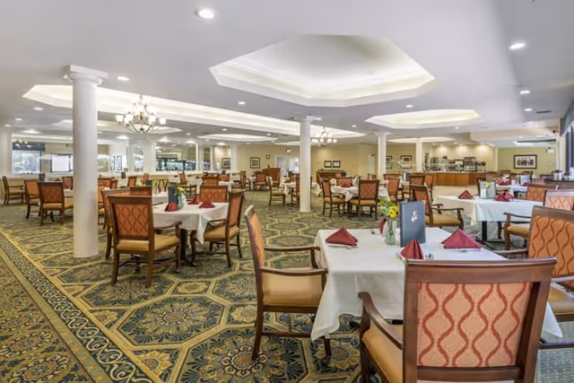 Spacious dining room with multiple tables covered in white tablecloths, each set with folded red napkins and menus. The room features patterned carpet, beige walls, white columns, and recessed ceiling lights with chandeliers. Large windows and mirrors line the walls, creating a bright and open atmosphere.
