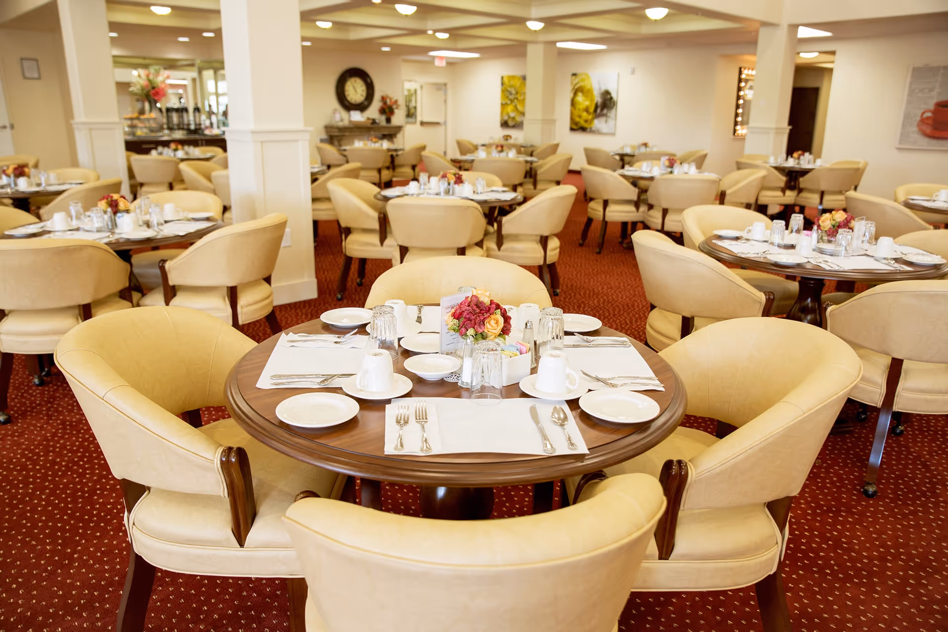 A spacious dining room with round wooden tables set for meals, each surrounded by beige upholstered chairs. The tables have white plates, cups, silverware, and small floral centerpieces. The room has a warm ambiance with carpeted floors and soft lighting.