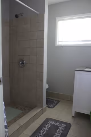 A bathroom with a tiled walk-in shower on the left, a window with blinds on the right wall, a white vanity cabinet, and two gray patterned bath mats on the tiled floor.