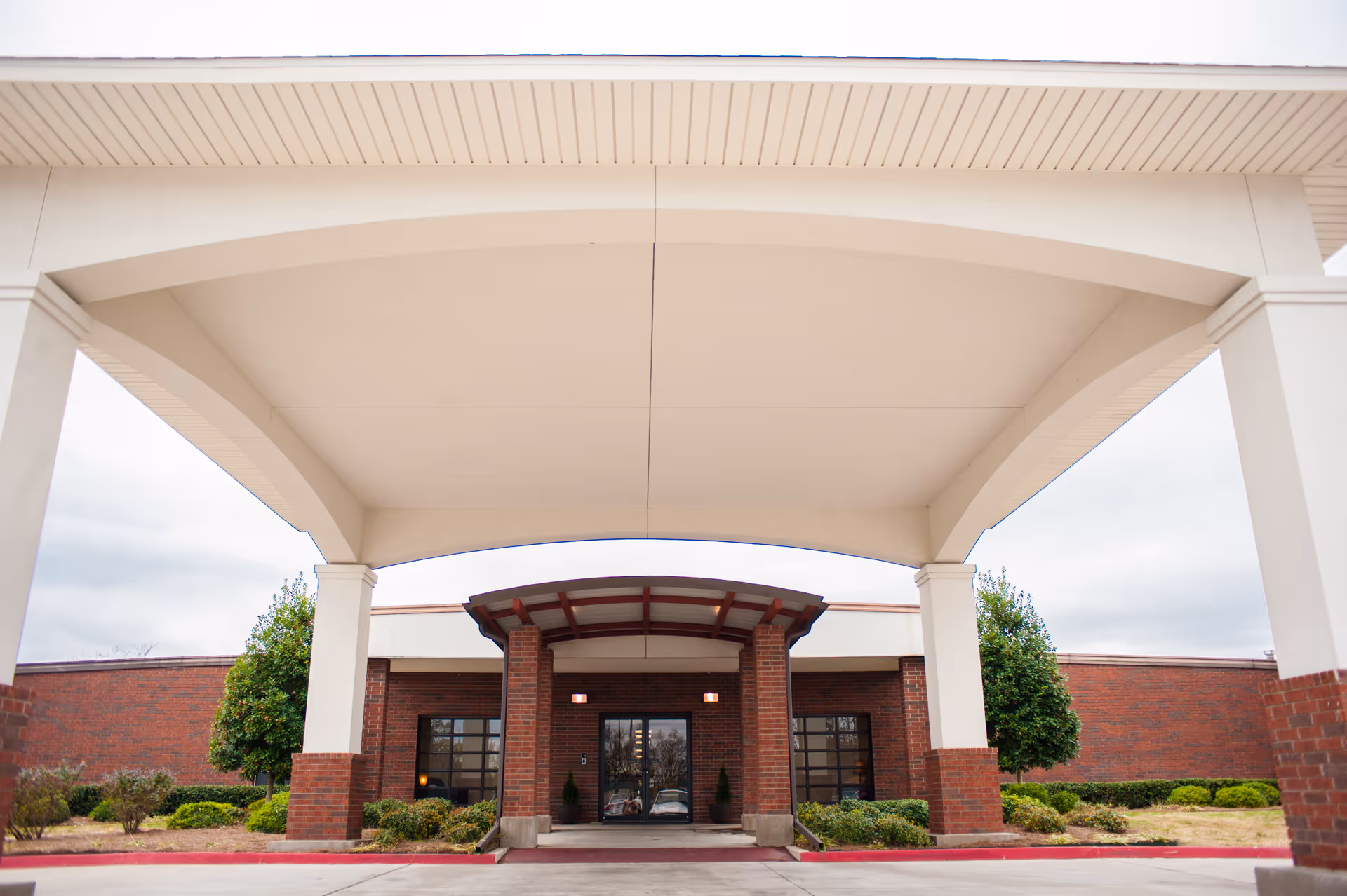 Entrance of Riverview Care Center showing a covered drop-off area with large white pillars and a brick facade. There are shrubs and small trees on either side of the entrance under a cloudy sky.