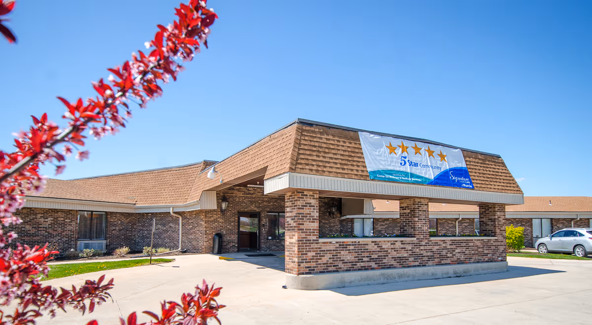 Single-story brick senior care building with a covered entrance and a large '5 Star Community' banner, set against a clear blue sky and framed by red flowering branches.