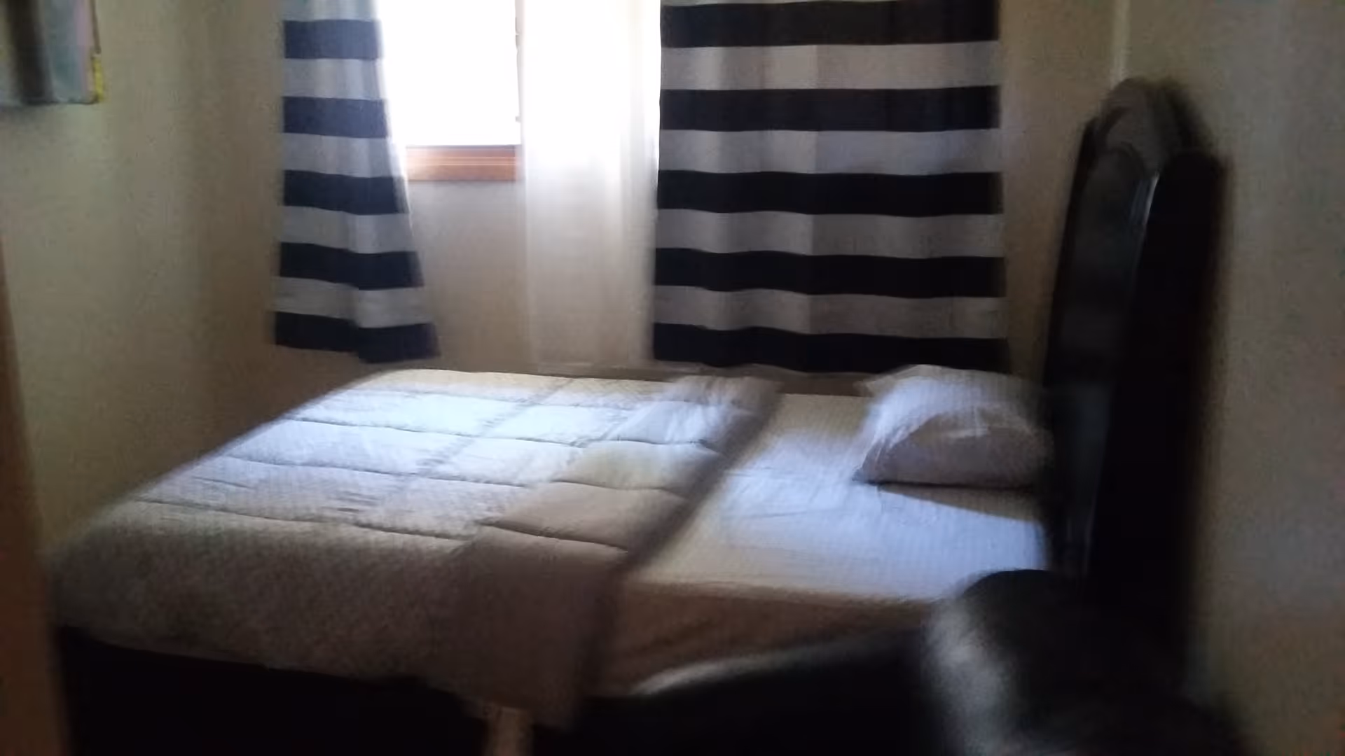 A small bedroom with a bed covered by a light-colored quilt and a single pillow. The bed has a dark wooden headboard. Behind the bed is a window with black and white striped curtains partially drawn, allowing some light into the room. The walls are light-colored, and a dark chair is partially visible in the foreground.