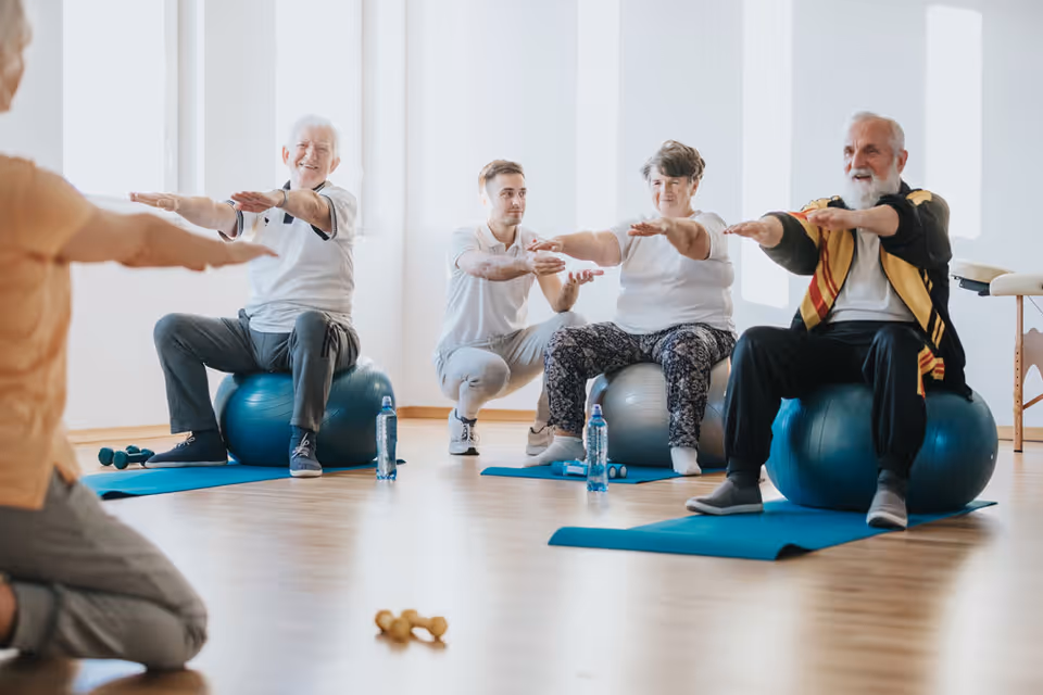 A group of elderly people sitting on exercise balls in a bright room, participating in a guided fitness session with a young instructor kneeling in front of them. They are extending their arms forward while seated on blue mats with water bottles and small dumbbells nearby.