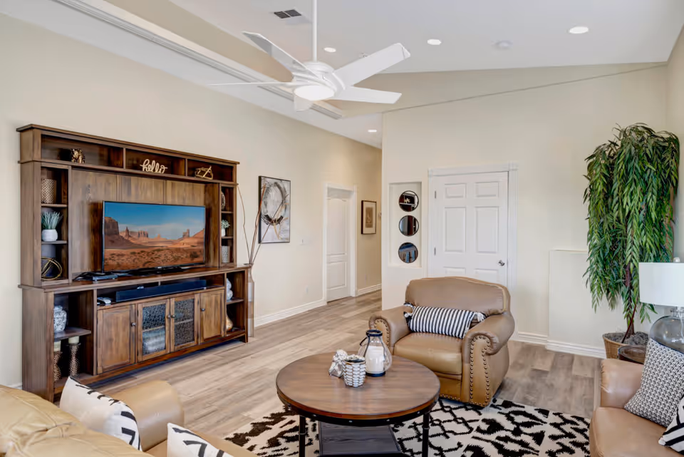 A cozy living room with beige leather armchairs and a sofa, a round wooden coffee table with decorative items, a large wooden entertainment center with a flat-screen TV displaying a desert landscape, a tall green plant in the corner, and light-colored walls and flooring.