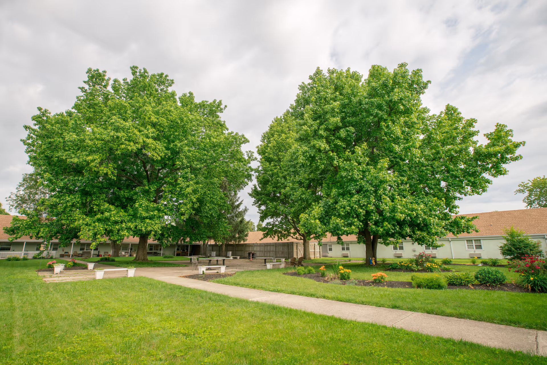 A well-maintained outdoor garden area at McNaughten Pointe featuring lush green grass, large leafy trees, flower beds with colorful blooms, concrete pathways, and several white benches arranged around the garden space. The background shows a single-story building with a brown roof and multiple windows.