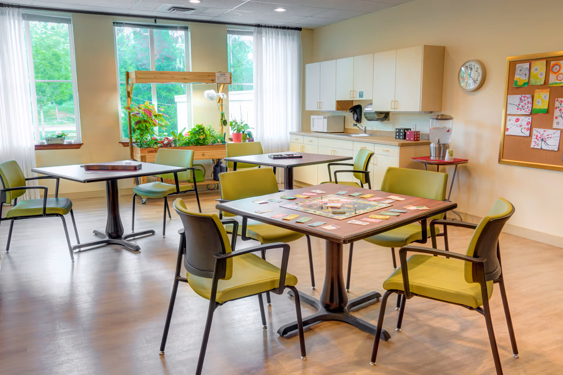 A bright communal activity room with green chairs around tables, a board game on the central table, a kitchenette, and plants by large windows.