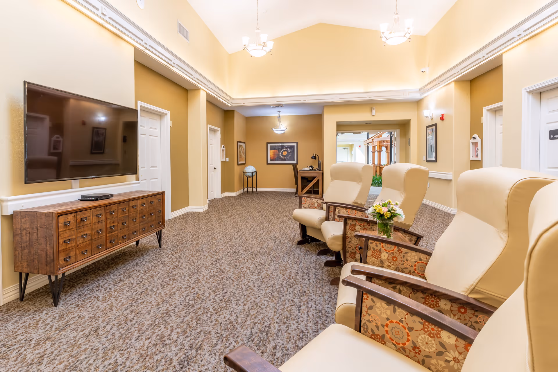 A well-lit common area in a senior living facility with beige walls and carpeted floor. There are several high-back armchairs with floral patterned armrests arranged facing a large flat-screen TV mounted on the wall above a wooden cabinet with multiple drawers. The room has multiple doors, framed pictures on the walls, and a small table with a globe and a desk with a lamp in the background. Ceiling lights and wall sconces provide illumination.