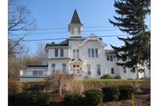 Large white multi-story building with a pointed tower, surrounded by trees and bushes under a clear blue sky.