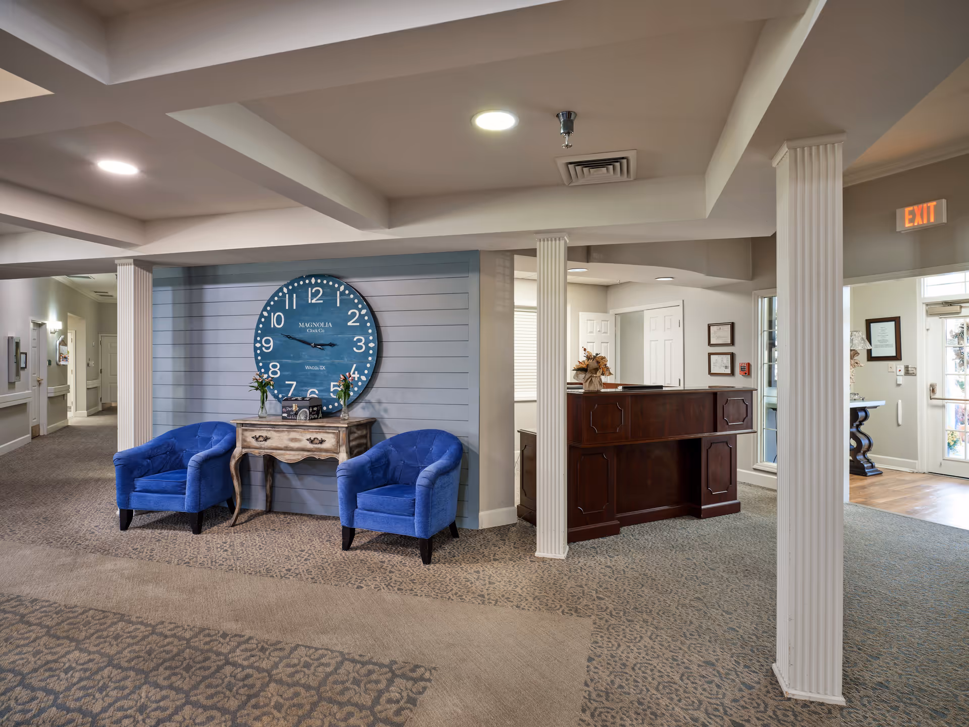 Interior view of a senior living facility lobby area with two blue armchairs flanking a small wooden table beneath a large wall clock. To the right is a dark wooden reception desk, and in the background there is an exit door with glass panels. The space is carpeted and has white columns and ceiling beams.