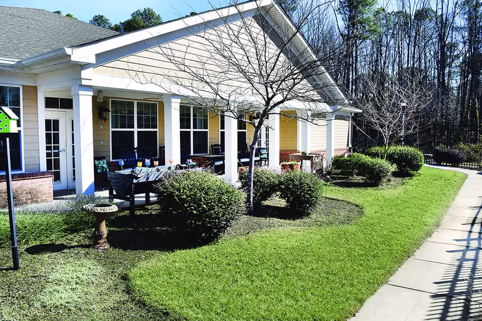Outdoor patio area of a senior living facility with a covered porch, several chairs, a table with colorful decorations, neatly trimmed bushes, a birdbath, and a paved walkway. Leafless trees and a wooded area are visible in the background.