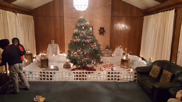 Decorated Christmas tree with presents and holiday displays in a wood-paneled common room with sofas and curtains.