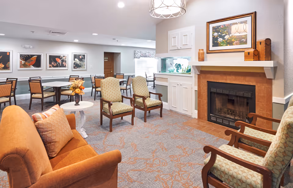 A cozy senior living facility common area with a fireplace, an aquarium built into a white cabinet, several cushioned armchairs, a small round table with a flower arrangement, and dining tables with chairs in the background. The walls are decorated with framed artwork and the room is well-lit with ceiling lights.