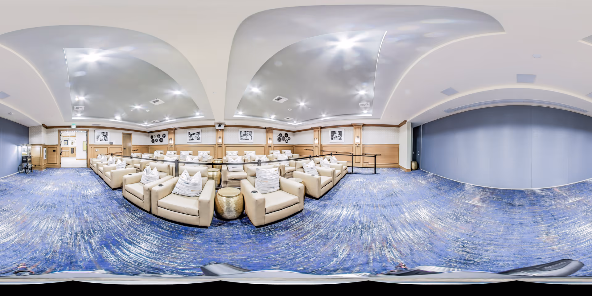 A spacious and well-lit common area with multiple beige armchairs arranged in rows, each with a white pillow. The room features blue carpeting, wooden paneling on the walls, and framed black and white photographs. The ceiling has recessed lighting and a modern design with curved elements.