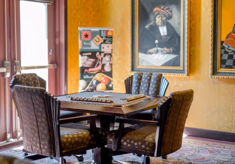A cozy game room with a square wooden table set for a tile-based game, surrounded by four cushioned chairs with patterned upholstery. The room has yellow patterned walls adorned with framed paintings, and a banner in the background advertising word games and billiards. Large glass doors allow natural light to enter the space.