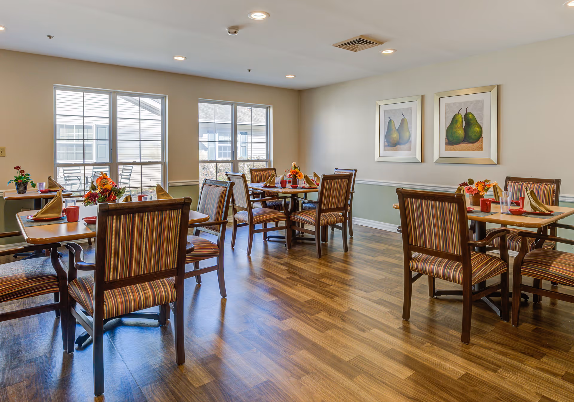 A bright dining room with several wooden tables and striped cushioned chairs. Each table is set with folded napkins, red cups, and small floral centerpieces. Two framed paintings of pears hang on the wall, and large windows allow natural light to fill the room.