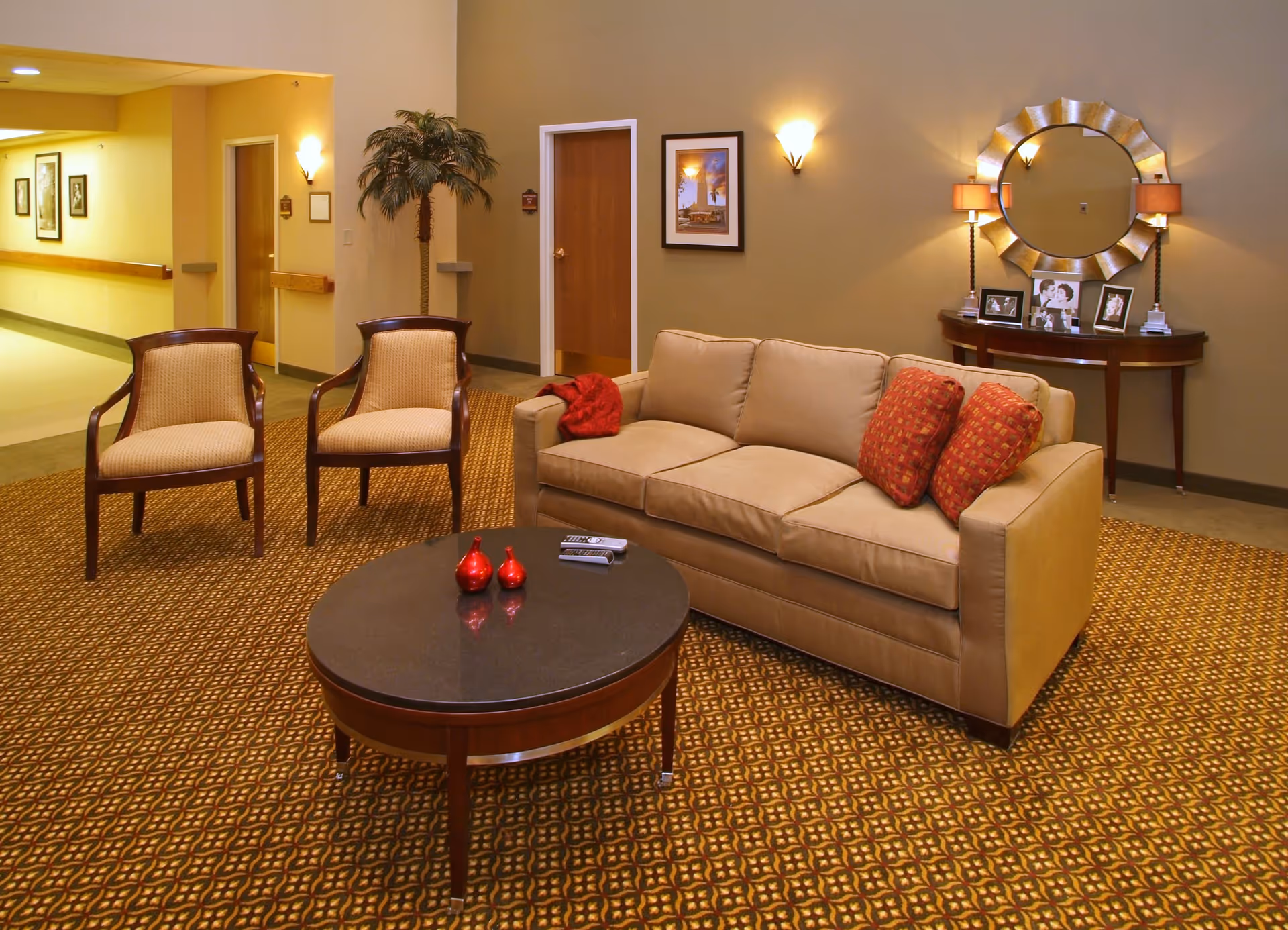 A cozy seating area in a senior living community featuring a beige sofa with two red patterned cushions and a red throw, two wooden chairs with beige upholstery, and a round coffee table with two red decorative items and two remote controls. Behind the sofa is a console table with framed photos, two lamps, and a large decorative round mirror on the wall. The room has warm lighting and patterned carpet, with a hallway and doors visible in the background.