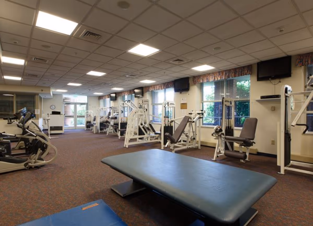 Large fitness room with exercise machines, weight equipment, and a central padded mat under a drop ceiling.
