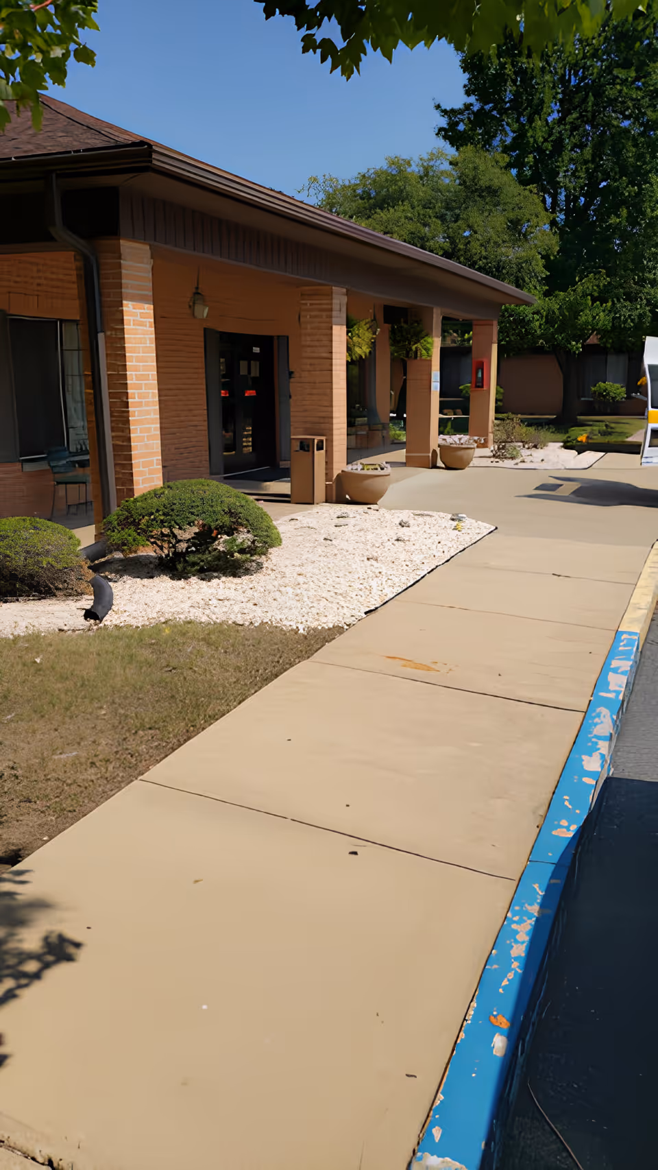 Exterior view of New Castle Health & Rehabilitation Center showing a brick building with a covered entrance, a concrete sidewalk, landscaped bushes, and a clear blue sky.