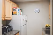 A small kitchen with wooden cabinets, a white refrigerator with cereal boxes on top, a microwave oven, a toaster oven, a clock on the wall, and a bowl of fruit on the counter.
