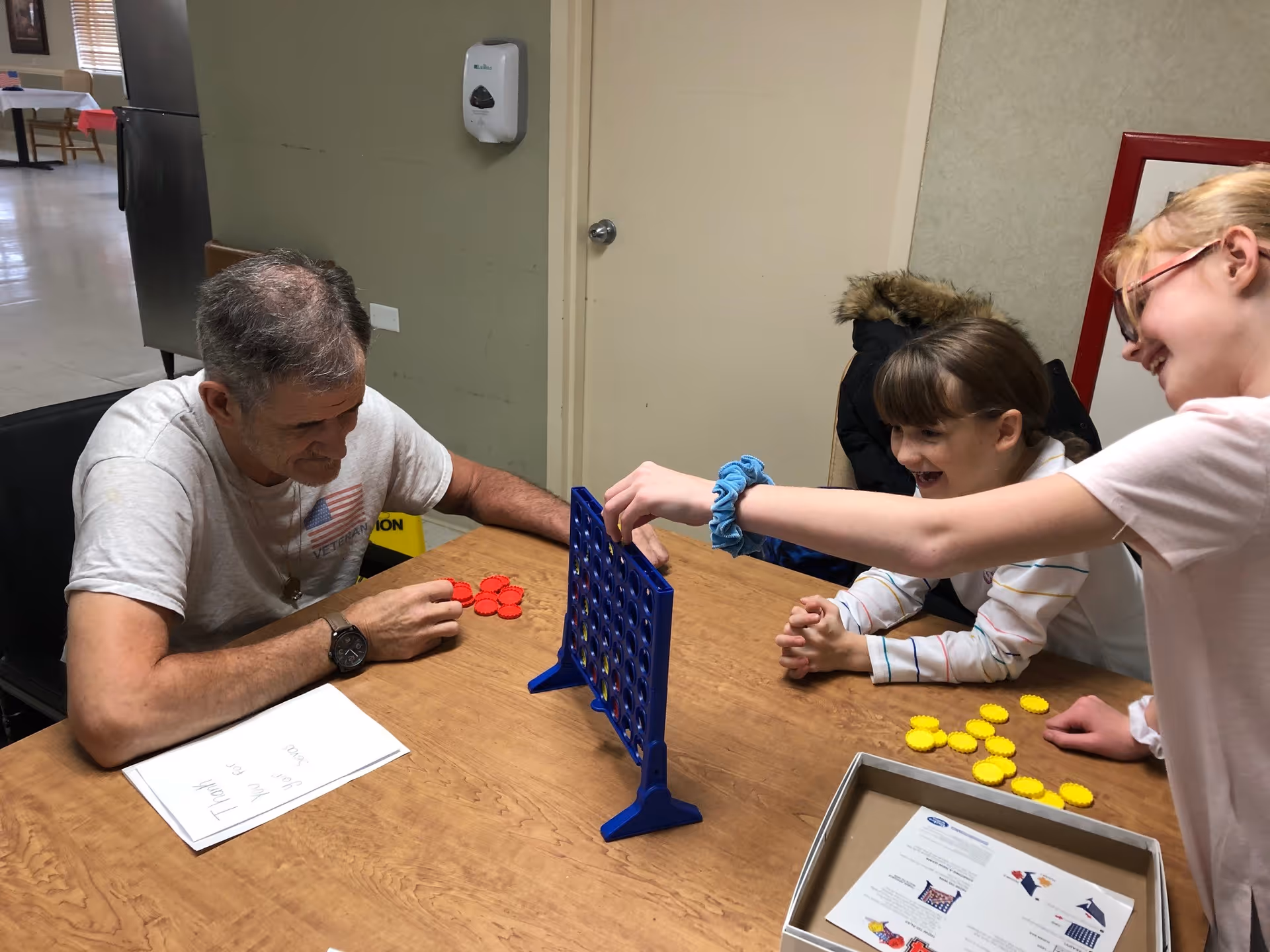 An elderly man and two young girls are sitting around a wooden table playing a game of Connect Four. The man holds red game pieces while one girl is placing a yellow piece into the blue game grid. They are smiling and appear to be enjoying the activity. The setting looks like an indoor common area with a hand sanitizer dispenser on the wall and some papers on the table.