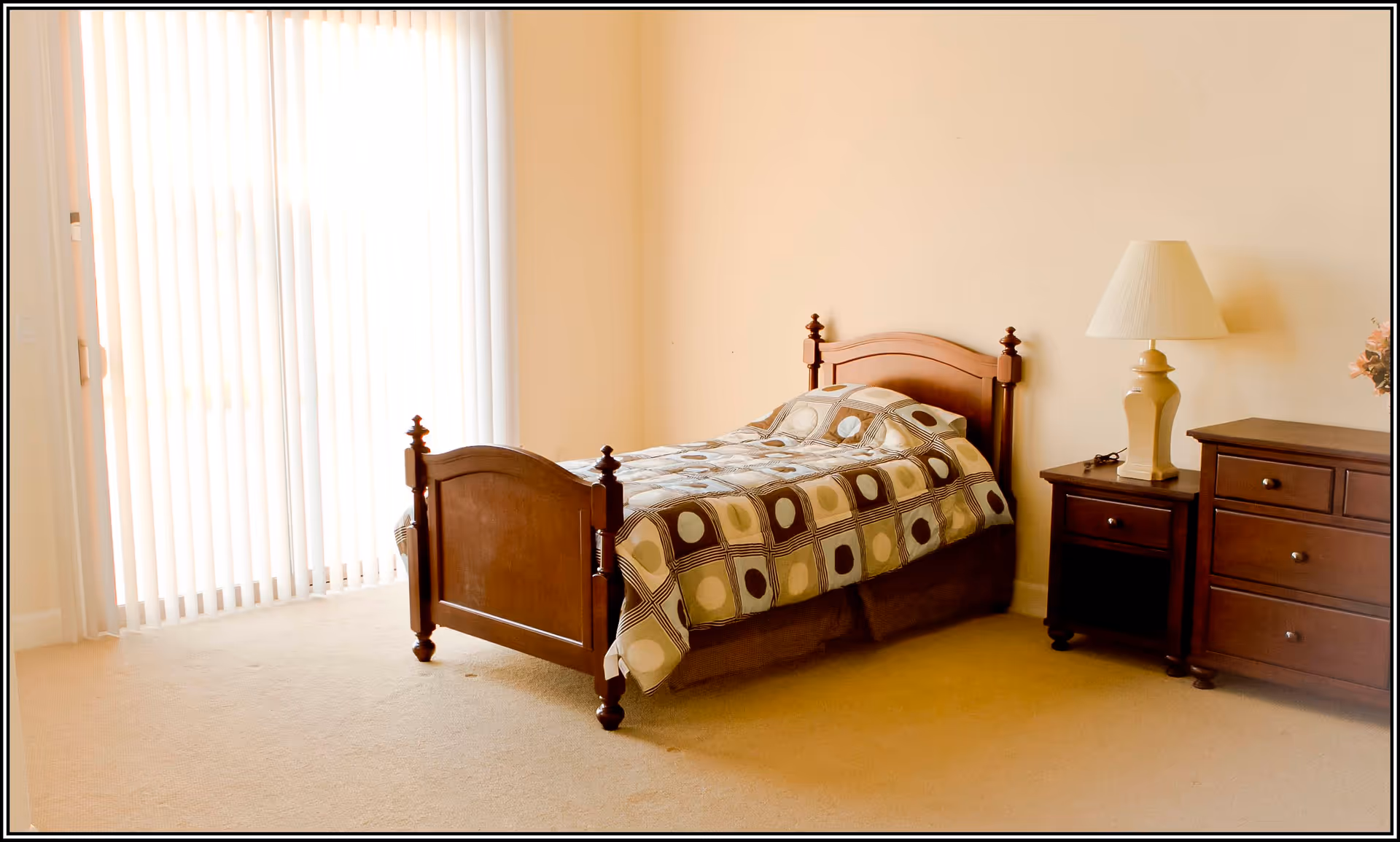 A simple bedroom with a single wooden bed covered with a patterned bedspread, a wooden nightstand with a lamp, and a wooden dresser. The room has beige walls and carpet, and a large window with vertical blinds letting in natural light.