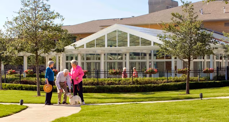 Three elderly people standing on a paved pathway in a grassy outdoor area with trees and flowers, one person is bending down to pet a small white dog. In the background, there is a large building with a glass-covered entrance and more people walking inside the fenced garden area.