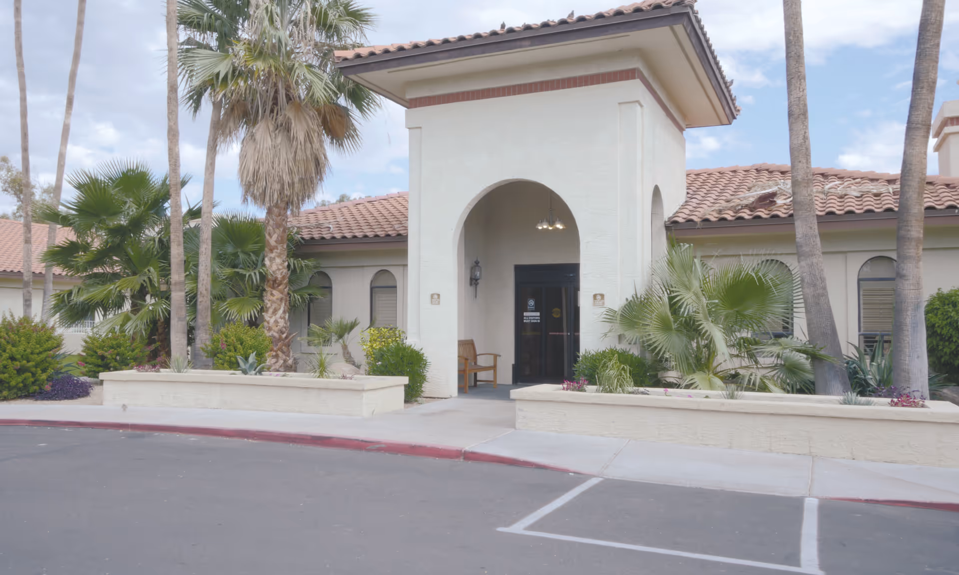 Entrance of a building with a covered archway, surrounded by palm trees and various green plants, with a bench near the door and a tiled roof under a partly cloudy sky.