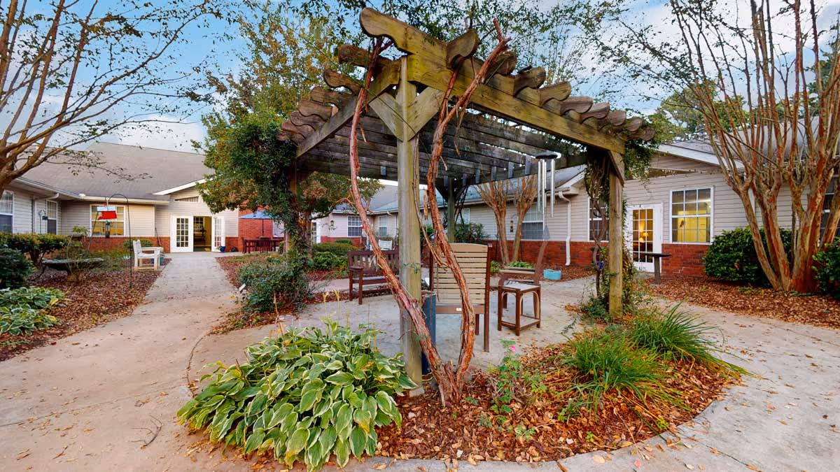 Outdoor courtyard area at Gaines Park Senior Living featuring a wooden pergola with climbing plants, surrounded by benches and chairs. The courtyard is landscaped with green plants and trees, with pathways leading to single-story residential buildings in the background under a clear sky.